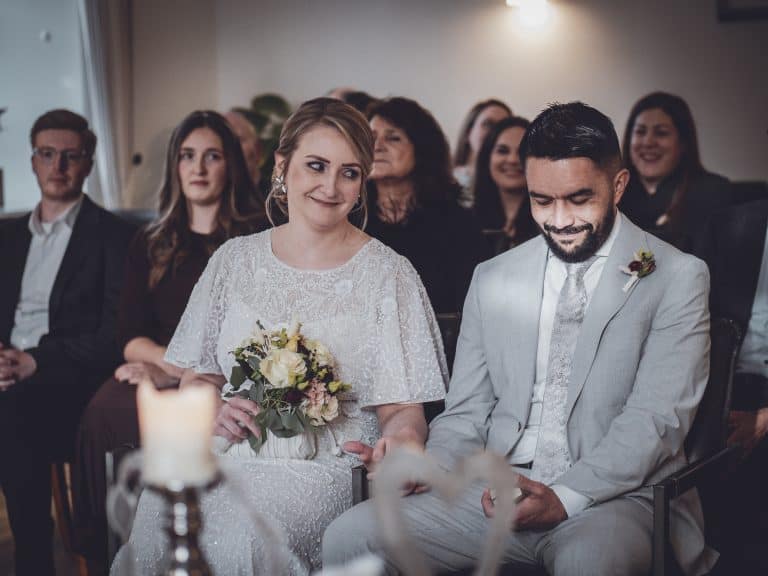 The bride holds a bouquet, smiling softly, while the groom looks down happily, surrounded by loved ones in a nostalgic, classic setting.