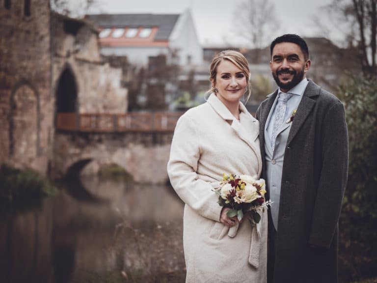 The couple stands close by a serene river, dressed elegantly for a special occasion, with a historic bridge and rustic buildings in the background, ev.