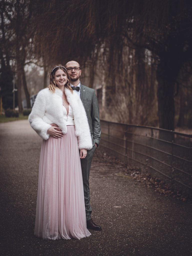 The couple stands close together on a pathway surrounded by tall trees, dressed elegantly in retro-inspired attire, radiating love and timeless charm.